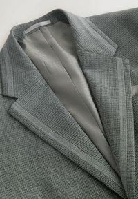 Sacou gri cu un model carouri texturat, având o căptușeală netedă din satin gri și reveruri cu crestături. Detalii de finisaj vizibile la margini.