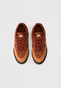 Nike SB kedas ķieģeļu brūnā zamšā ar tumšāku brūnu purngalu. Iezīmes ietver plakanās šņorītes, baltu logo un melnu gumijas zoli.