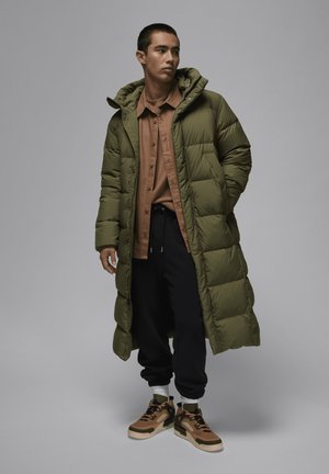 Lang olivengrøn dunjakke med hætte, brun skjorte med knapper, sorte sweatpants og multicolor sneakers med strukturerede detaljer.