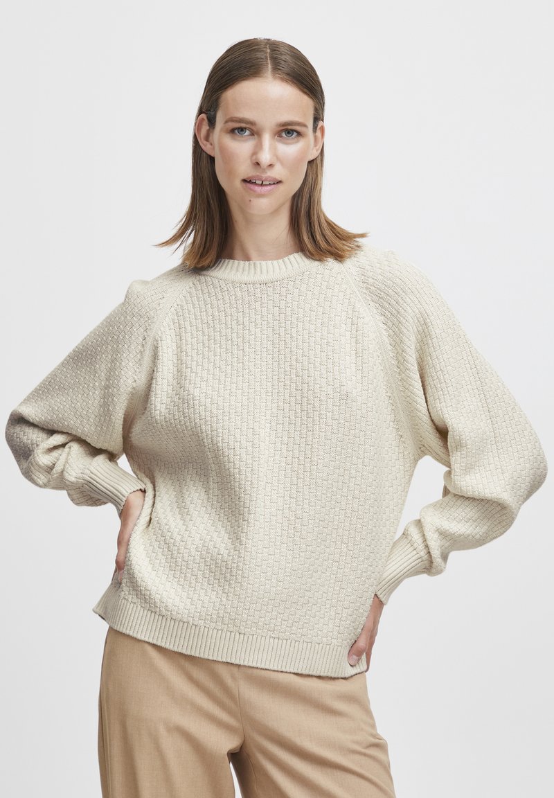 Pull en crème texturé avec un col et des poignets côtelés. Présente une coupe décontractée et un motif distinctif en forme de gaufre sur l'ensemble.