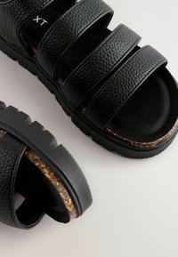 Schwarze Sandalen mit drei strukturierten Riemen, einem weichen gepolsterten Fußbett und einer robusten Gummisohle mit geriffeltem Rand und Korkakzent.