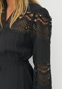 Mustmust, mille keerukate pitsdetailidega varrukatel, tekstureeritud kangas, elastne vöökoht ja mugav lõige.