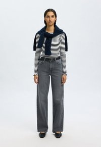 Csíkos hosszú ujjú felső, tengerészkék részletekkel díszített kötött sál, magasderekú, széles szárú szürke farmernadrág, fekete öv és fekete lapos cipő egyszínű háttér előtt.