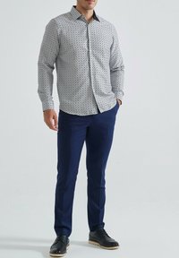 Homme portant une chemise blanche à motifs, un pantalon bleu marine et des chaussures de ville noires, se tenant avec une main dans la poche.