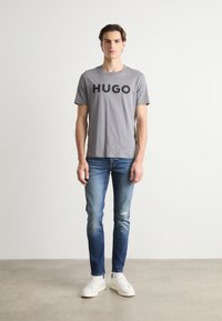 Tricou gri din bumbac cu text negru îndrăzneț "HUGO", asortat cu blugi decolorați și adidași albi, design minimalist pe un fundal neutru.