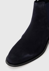 Dunkelmarineblaue Wildleder-Chucka-Stiefel mit runder Zehenpartie und schwarzer Sohle auf einem weißen Hintergrund.
