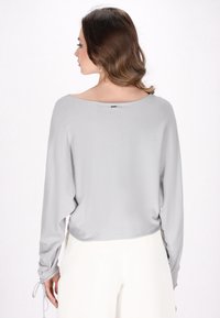 Top a maniche lunghe grigio chiaro con vestibilità morbida, scollatura bateau, polsini arricciati e texture liscia, abbinato a pantaloni bianchi.