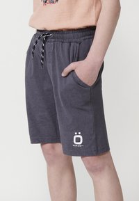 Sivé bavlnené šortky s elastickým pásom a čierno-bielym šnúrkovým zapínaním. Obsahujú bočné vrecká a biele logo na spodnej časti nohy.