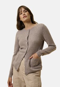Světle hnědý cardigan vyrobený z měkkého materiálu, s kulatým výstřihem, předními knoflíky, dvěma kapsami a žebrovanými manžetami.