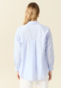 Camicia azzurro chiaro con righe bianche verticali, dotata di colletto, maniche lunghe e orlo leggermente curvo sul retro.