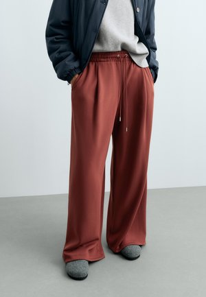Personne portant un pantalon ample couleur rouille, des chaussons gris, un pull gris et une veste sombre avec les mains dans les poches sur un sol uni.
