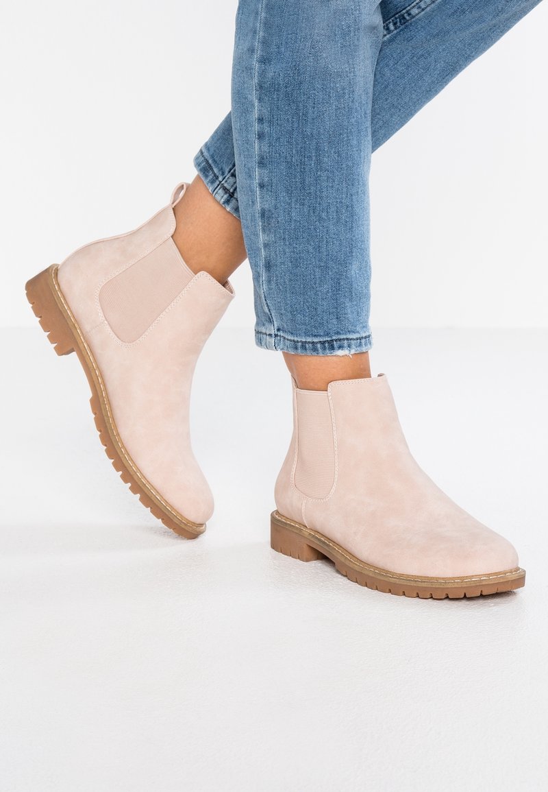 Botas de tobillo de ante rosa claro con paneles laterales elásticos y suela de goma texturizada. Llevadas con jeans azules con vuelta sobre un fondo blanco.