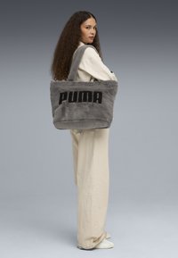 Borsa a tracolla grigia in peluche con logo "PUMA" nero, texture morbida, forma rettangolare e due manici robusti, indossata sulla spalla.