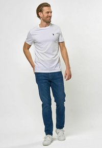 Fehér pamut póló lekerekített nyakkivágással, bal mellrészen egy kis tengerész kékkel logóval, sötétkék farmernadrággal és fehér sportcipővel párosítva.