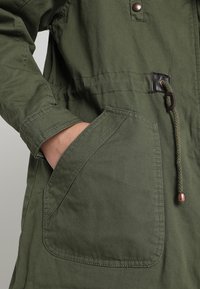 Main reposant dans la poche avant d'une veste vert olive avec cordon de serrage et fermeture à bouton-pression.
