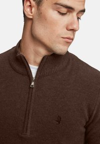 Suéter marrón de punto con cuello alto y media cremallera. Presenta un cuello acanalado y un sutil detalle de logo en el pecho. La textura parece suave.