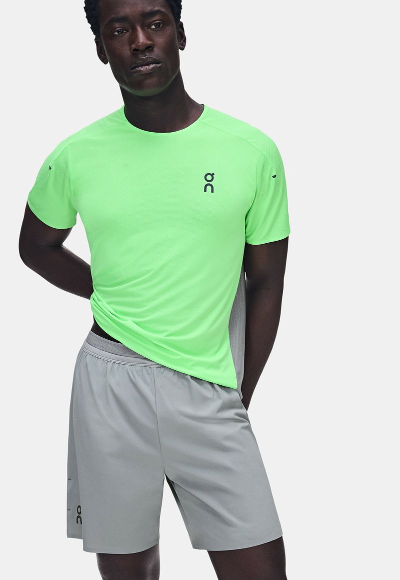 Homme portant un t-shirt de sport vert vif à manches courtes et un short gris clair, debout avec une main derrière le dos.