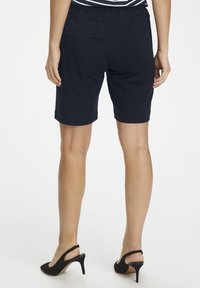 Donna che indossa pantaloni corti neri al ginocchio e sandali con tacco a slitta neri, in piedi contro uno sfondo bianco, vista da dietro.