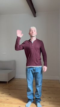 Jeune homme aux cheveux blonds portant une chemise bordeaux et un jean, fait un signe de la main droite dans une pièce peu meublée avec un sol en bois.