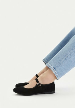 Füße in hellblauen Jeans, die schwarze Mary Jane Ballerinas aus Wildleder mit einem dünnen Riemen und einer kleinen Schnalle tragen, vor einem schlichten weißen Hintergrund gezeigt.