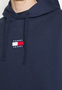 Felpa con cappuccio blu navy realizzata in tessuto morbido, caratterizzata da una patch rettangolare con logo bianco, rosso e blu con il testo "TOMMY JEANS."