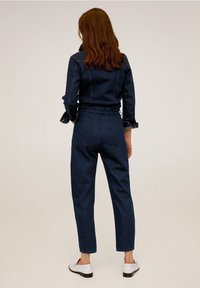 Jumpsuit de mezclilla azul oscuro con un corpete ajustado, cintura elástica y piernas ajustadas. Presenta costuras clásicas y mangas con puños.