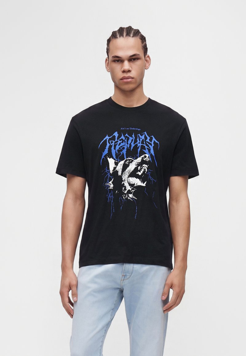 Νέος άνδρας με πλεξούδες, που φοράει μαύρο T-shirt με γραφικό ενός σκύλου που γρυλίζει και μπλε κείμενο, συνδυασμένο με ανοιχτό μπλε τζιν, στέκεται μπροστά σε λευκό φόντο.