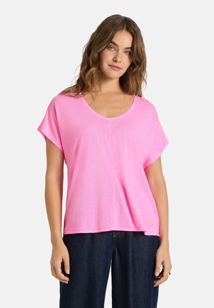 Frau mit welligem braunem Haar, die ein lockeres pinkfarbenes Kurzarmshirt und dunkelblaue Jeans trägt, steht vor einem schlichten weißen Hintergrund.