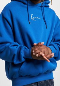 Sudadera con capucha azul con cordón ajustable, puños acanalados y un logo blanco bordado en el pecho. Tela suave con un corte relajado.