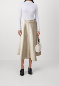 Beige læder midi nederdel med en udvidet design og sømdetaljer, parret med en hvid langærmet top og sorte hæle mules. Holder en lille hvid taske.