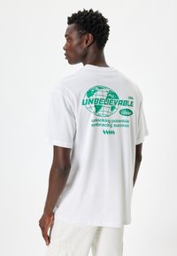 Hvid bomulds T-shirt med grøn grafisk print med globusdesign og tekst på ryggen. Korte ærmer, afslappet pasform og rund hals.
