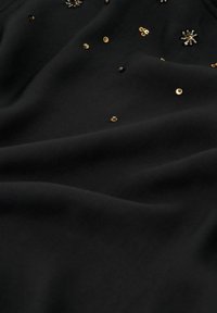 Zwarte stof met een gladde textuur, voorzien van gouden pailletten en decoratieve elementen, die een subtiel contrast en ingewikkelde visuele details creëren.