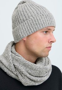 Berretto lavorato a maglia grigio con una texture a coste e un piccolo pompon sulla parte superiore, abbinato a una sciarpa in maglia grigia, che offre un aspetto morbido e accogliente.