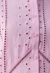 Blusa rosa ricamata con punti decorativi, motivi triangolari e floreali, e piccoli bottoni lungo la parte anteriore.