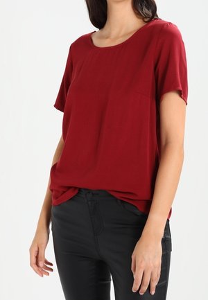 Kvinde iført en kortærmet burgundy top, stoppet ned i sorte tætsiddende bukser, stående mod en ensfarvet hvid baggrund.