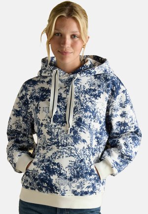 Young woman wearing a blue and white patterned hoodie with forest and animal designs, hands in pockets, smiling softly, light background.