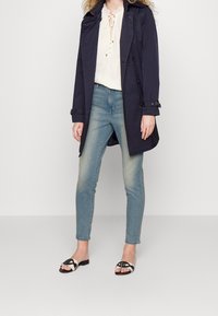 Marinblå trenchcoat med dubbelknäppning över en vit spetsblus, tillsammans med ljusblå skinny jeans och sandaler i svart och vitt.