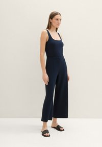 Mono de rayas azul marino con pantalones de pierna ancha, cuerpo ajustado y escote cuadrado, combinado con sandalias deslizantes negras sobre un fondo neutro.