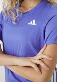 Person med lockigt blont hår som bär en lila sporttröja och har armarna korsade, vilket visar en vit geometrisk logotyp på bröstet.