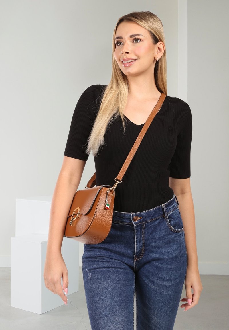 Bruine leren crossbody tas met een ronde vorm, een zilveren sluiting en een kleurrijke label. Draag met een zwart geribbeld topje en blauwe jeans.