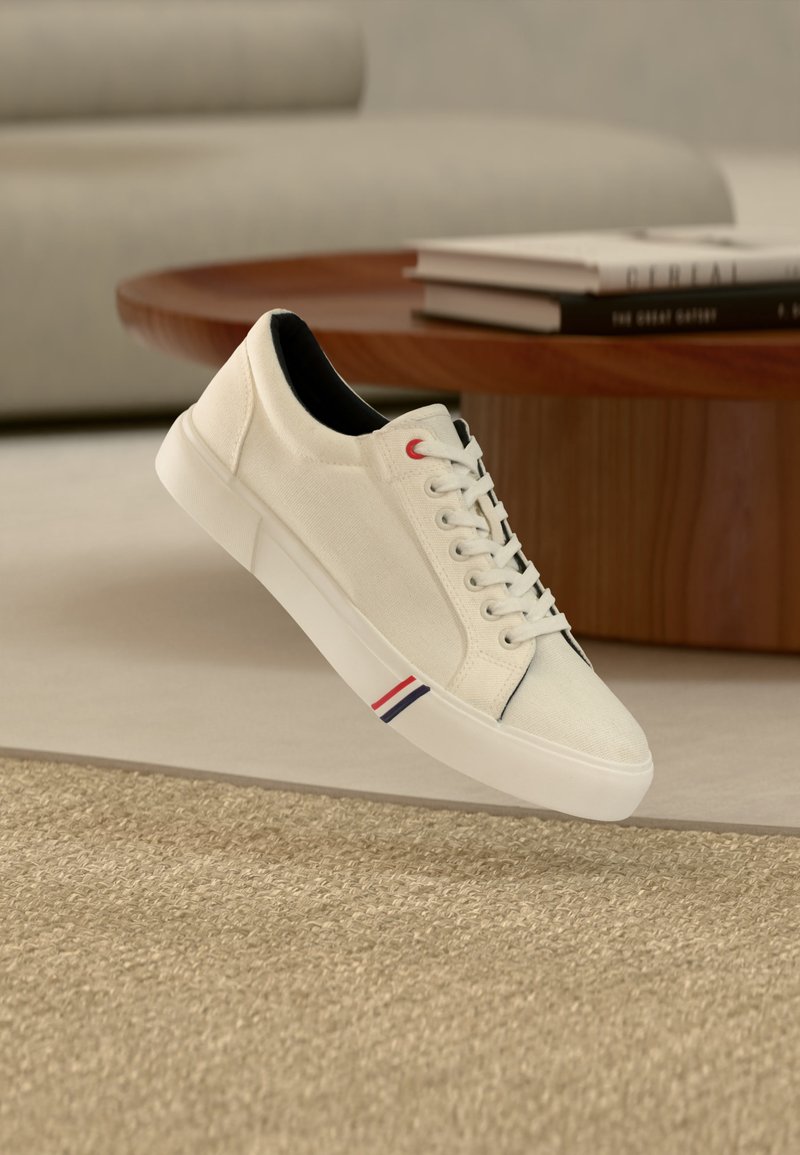 Λευκό low-top αθλητικό παπούτσι με κορδόνια, μαύρο εσωτερικό και κόκκινες, λευκές, μπλε ρίγες κοντά στη σόλα, τοποθετημένο σε εσωτερικό χώρο σε μπεζ χαλί κοντά σε ξύλινο τραπέζι.