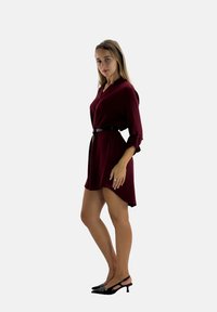 Robe bordeaux avec un col et une taille ceinturée, dotée de manches longues et d'un ourlet asymétrique. Portée avec des chaussures à talons moyens en vernis noir.