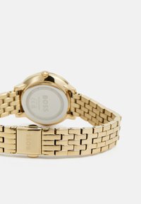 Montre-bracelet dorée avec un bracelet en maillon métallique, boîtier rond et surface lisse et brillante, dotée d'un fermoir et d'une gravure de marque sur le bracelet.