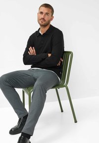 Polo a maniche lunghe nera, pantaloni grigi dalla texture, seduto su una sedia di metallo verde. Scarpe nere con superfici texture. Design semplice e moderno.