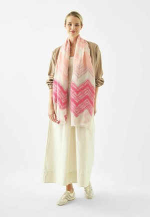 Woman wearing beige jacket, white dress, white sneakers, and a long pink and white chevron-patterned scarf standing against white background.