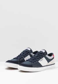 Námořnické plátěné tenisky s bílou mezipodešví, šedými a krémovými semišovými akcenty a červeným detailem na patě. Obsahují ploché tkaničky a štítek s logem na jazyku.