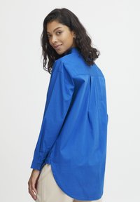 Blue button-up shirt with long sleeves, curved hem, back pleat detail, made of lightweight fabric. Worn over light-colored pants.