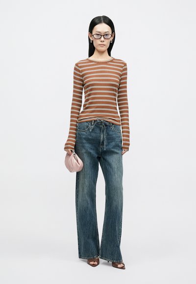 Top de manga comprida às riscas em castanho com linhas brancas, combinado com jeans azuis largos. Segurando uma bolsa rosa clara, usando sandálias castanhas com salto.