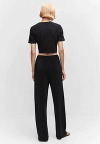 Personne aux cheveux blonds courts, vue de dos, portant un t-shirt noir court et un pantalon noir ample, sur un fond uni.