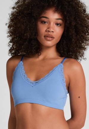 Femme aux cheveux bouclés portant un bralette côtelé bleu clair avec bordure en dentelle et bretelles réglables, posant devant un fond neutre.